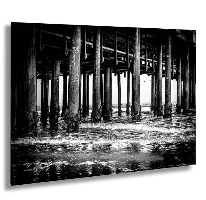 A black and white photograph of a pier with multiple wooden columns and beams, with water flowing under the structure.