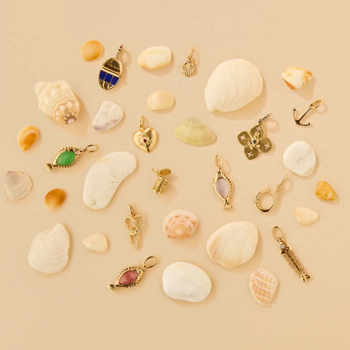 A flat-lay arrangement of various solid gold jewelry charms styled with white and pastel seashells on a solid beige background.