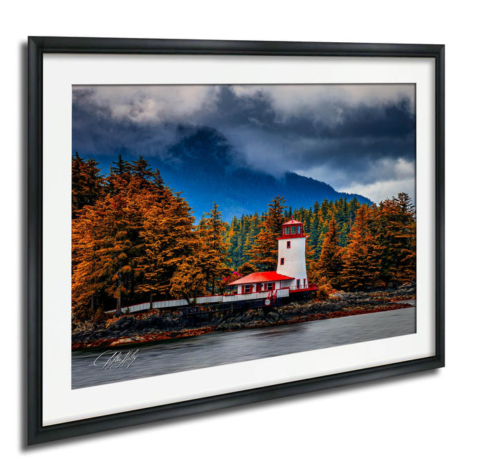 A framed photograph depicting a lighthouse surrounded by a forest with autumn-colored trees and a cloudy sky in the background.