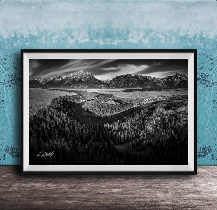 A black and white landscape photograph depicting a mountainous, forested area with a winding river or stream in the foreground.