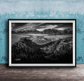 A black and white landscape photograph depicting a mountainous, forested area with a winding river or stream in the foreground.