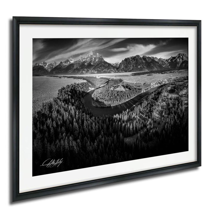 A black and white landscape photograph depicting a winding road surrounded by a forested area, with snow-capped mountains in the background.