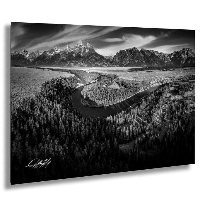 A black and white landscape photograph depicting a winding river surrounded by a forested area with snow-capped mountains in the background.