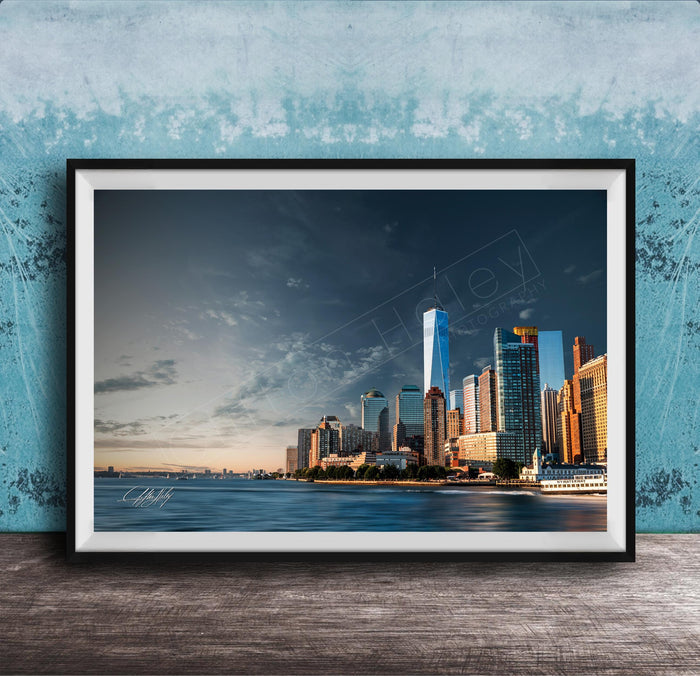 A framed photograph of the New York City skyline, with the One World Trade Center prominently visible, set against a blue sky with clouds.