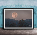 A framed photograph of a large, full moon in the night sky, with silhouetted trees and a blue sky in the background.