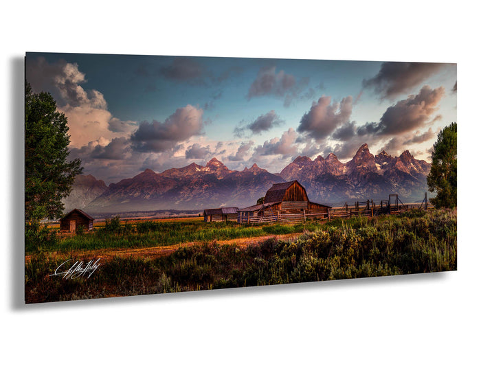 A picturesque rural landscape with a wooden barn, a small shed, and a dirt road leading to a mountain range in the distance. The sky is filled with clouds, and the sun is setting, casting a warm glow over the scene.