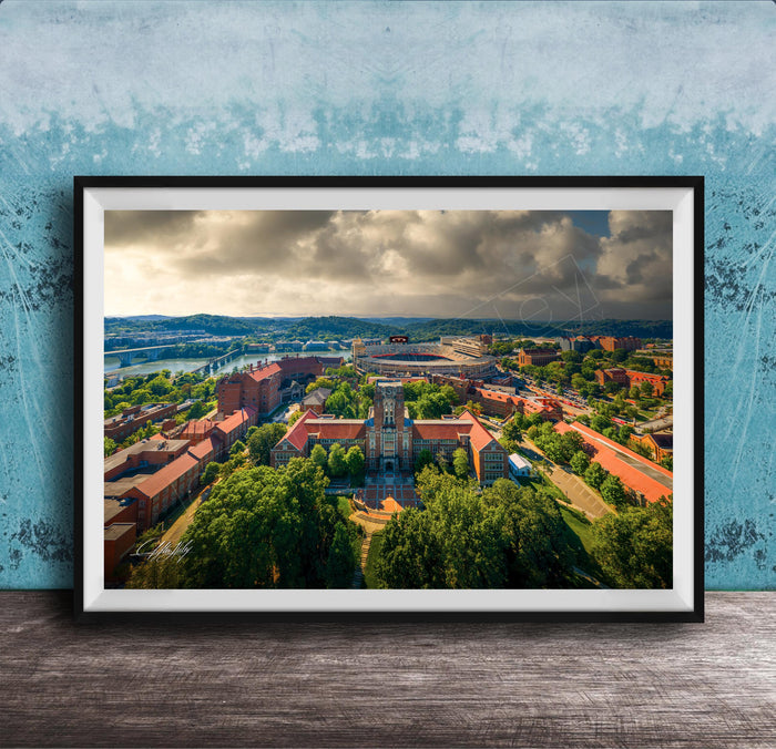 The image is a framed aerial view of a city with a mix of buildings, trees, and a river. The sky is cloudy, creating a dramatic backdrop for the cityscape.