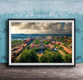The image is a framed aerial view of a city with a mix of buildings, trees, and a river. The sky is cloudy, creating a dramatic backdrop for the cityscape.