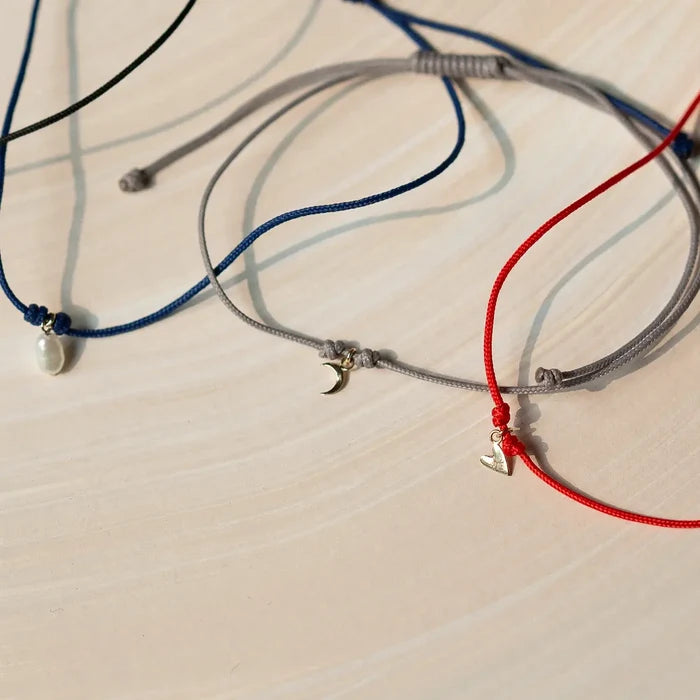 Three adjustable cord bracelets are neatly arranged on a light surface. The collection includes a grey, blue, and red bracelet from bluboho. Each bracelet showcases a small, simple charm: the grey and blue cords display a crescent moon charm, while the red cord features The Deep Love Contemplation Cord Bracelet adorned with a star charm in 10k yellow gold.