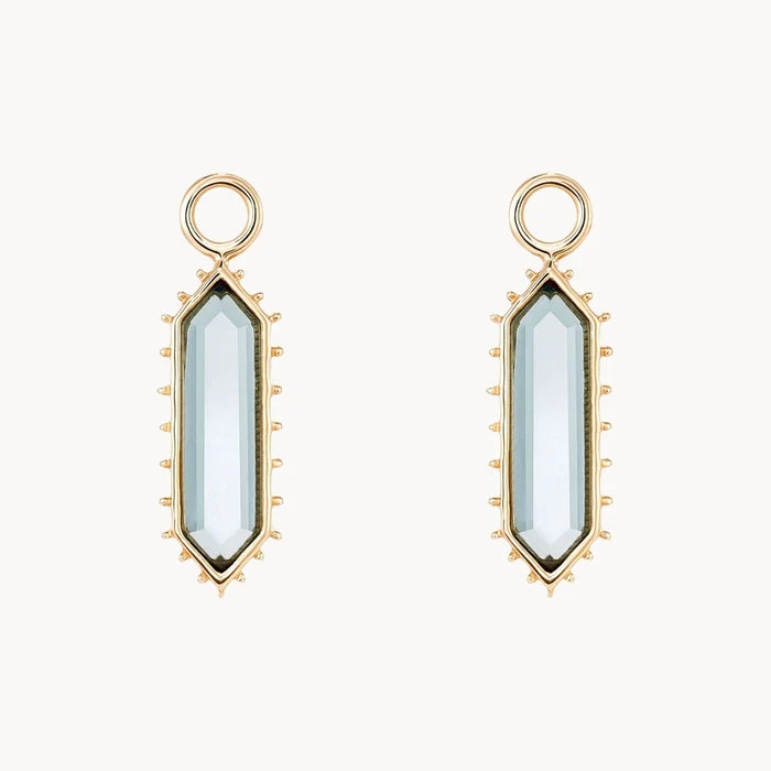 A pair of gold elongated hexagon earring charms with a london blue topaz stone surrounded by spiked edges, shown on a plain off-white background.