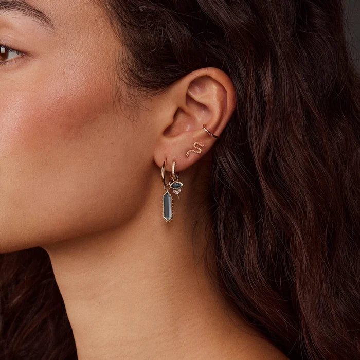 Close-up of a model's ear wearing multiple delicate solid gold earrings, including a small dangling earring with a london blue topaz rectangular stone.