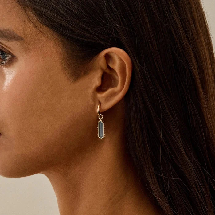 A close-up side profile of a woman wearing a delicate gold huggie hoop earring with a vertical, elongated oval london blue topaz stone in a bezel setting.
