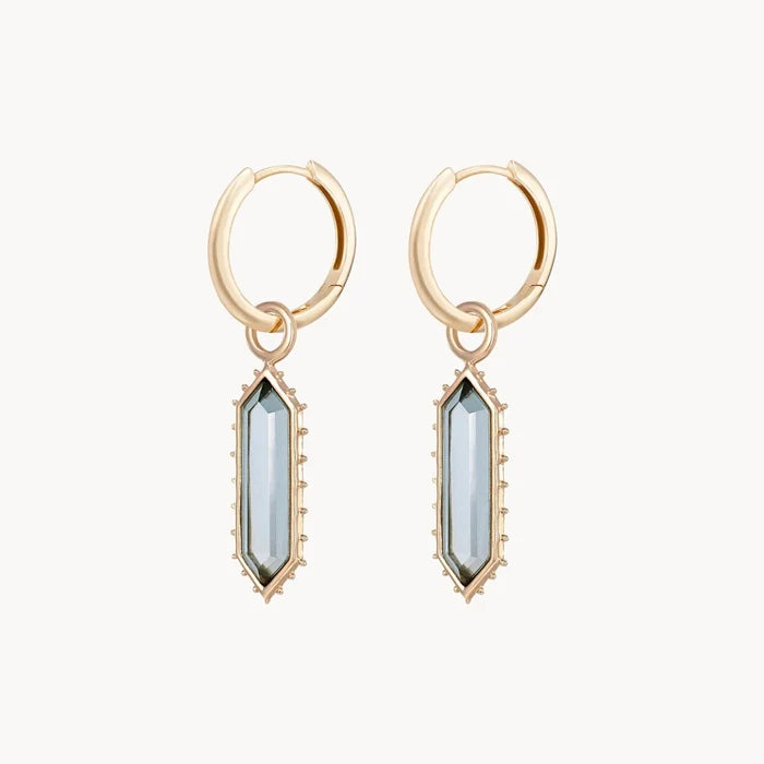 A side view of a pair of elongated hexagon earring charms with a london blue topaz stone surrounded by spiked edges, hung from a pair of gold hoop earrings, shown on a plain off-white background.