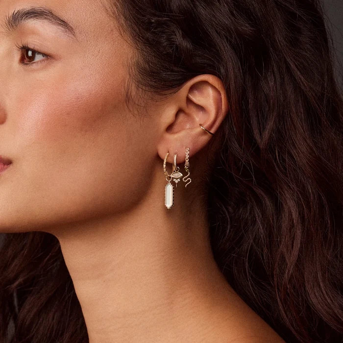 A close-up of a model's ear wearing a stack of solid gold hoops and earring charms, including a small snake charm and an evil eye pendant, all with natural diamonds and stones