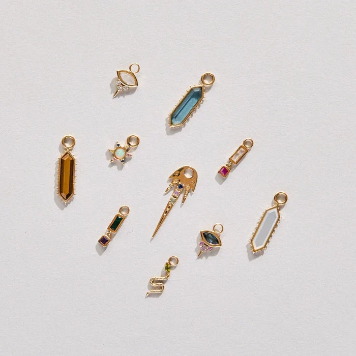 A set of nine solid gold earring charms arranged on a clean, off-white background. The charms feature various shapes including points, teardrops, and abstract designs, embellished with colourful stones such as blue, green, pink, and purple.
