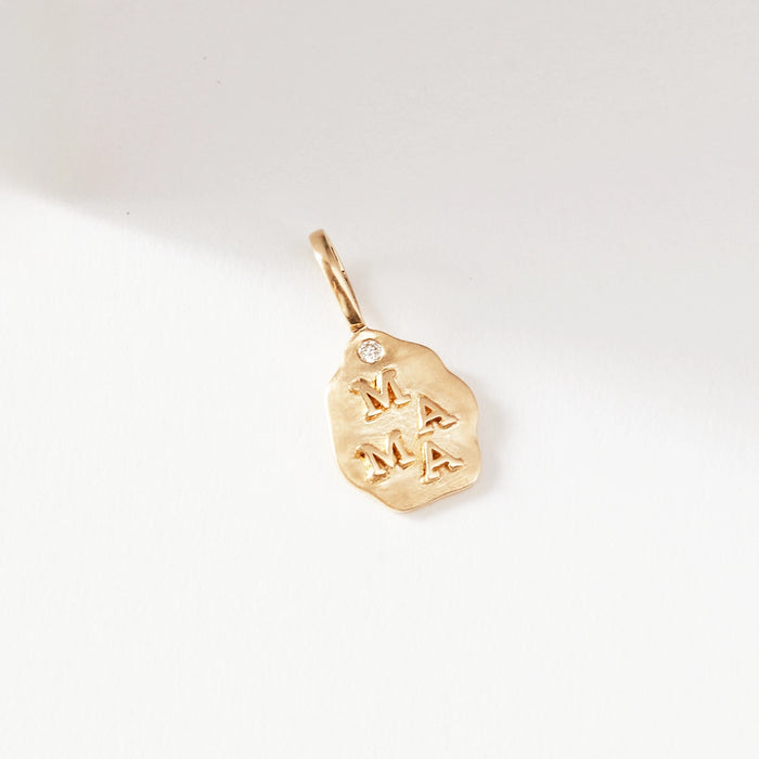 A close-up photograph of "the mama charm" in 10k yellow gold by bluboho. The charm features an irregular, nugget-like shape with a small diamond attached at the top. The word "MAMA" is whimsically engraved on its surface in an uneven style. The background is a plain off-white color.