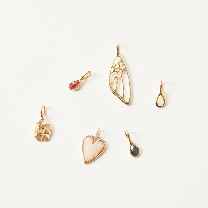 A collection of six assorted bluboho "the mama charm" 10k yellow gold pendants arranged on a white background. The pendants feature various shapes and designs: an abstract shape, a heart with a pearl-like center, a teardrop with a blue stone, a teardrop with a red stone, a wing-like shape, and a small loop.