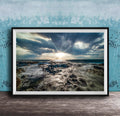 The image is a framed photograph of a rocky coastline with crashing waves, set against a backdrop of a cloudy sky and the sun peeking through the clouds.