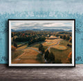The image is a framed aerial view of a golf course surrounded by trees and rolling hills, with a blue sky and clouds overhead.
