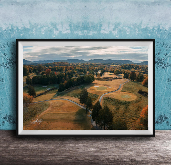 The image is a framed aerial view of a golf course surrounded by trees and rolling hills, with a blue sky and clouds overhead.