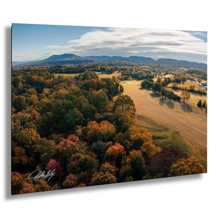 The image is a scenic aerial view of a countryside landscape with a mix of trees in autumn colors, rolling hills, and a distant mountain range under a blue sky with clouds.