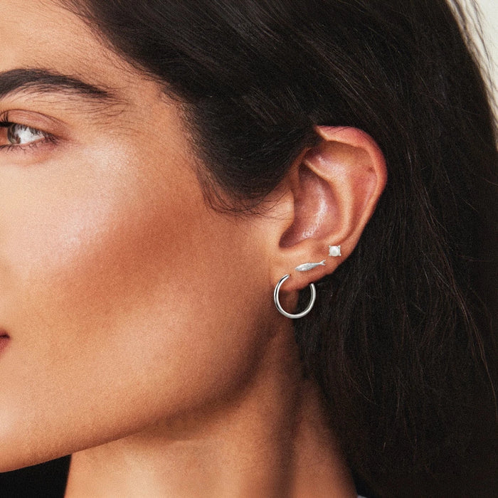 Close-up of a woman's ear wearing three silver earrings: a small pearl stud, a little fish stud, and a perfect everyday large hoop.