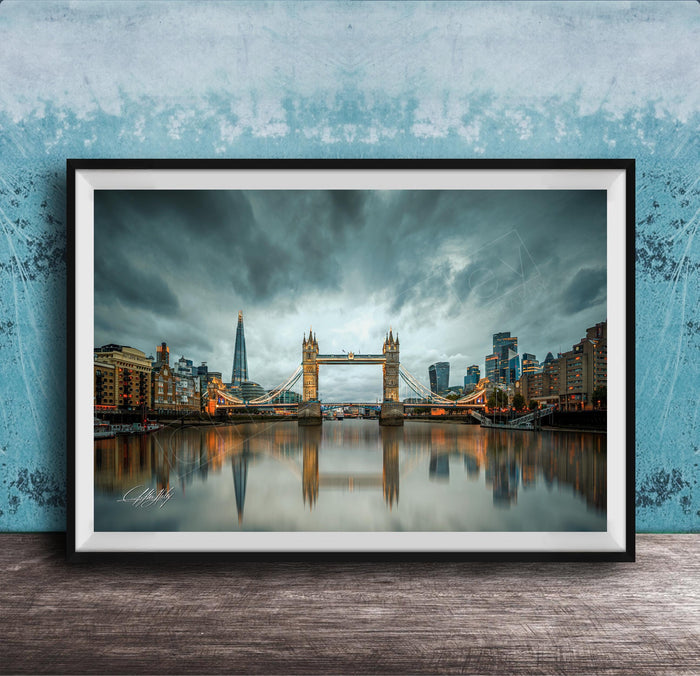 A framed photograph of the Tower Bridge in London, England, with the city's skyline reflected in the water below. The sky is filled with clouds, creating a dramatic atmosphere.
