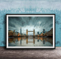 A framed photograph of the Tower Bridge in London, England, with the city's skyline reflected in the water below. The sky is filled with clouds, creating a dramatic atmosphere.