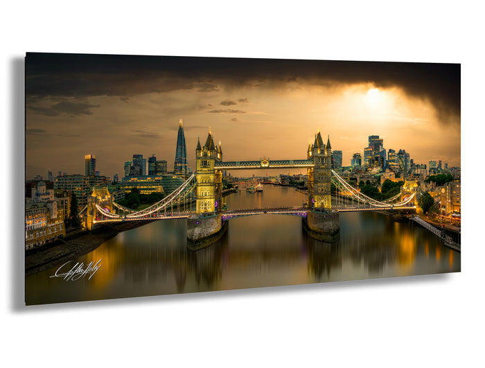 A panoramic view of the London skyline at night, featuring the iconic Tower Bridge spanning the River Thames. The bridge is illuminated with lights, creating a striking contrast against the dark sky. The cityscape is reflected in the calm waters of the river, adding to the overall beauty of the scene.