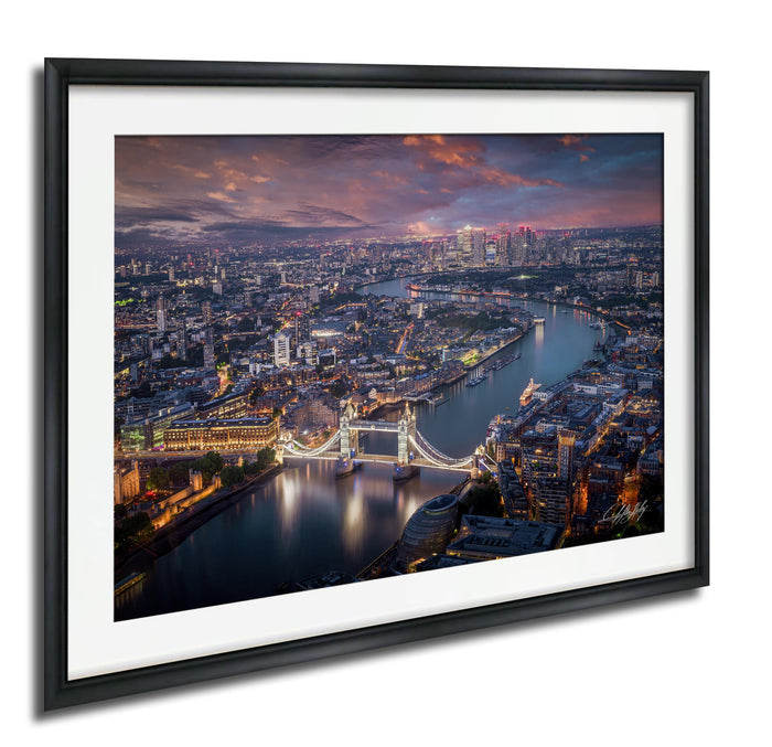 The image is a framed aerial view of a city at night, showcasing a river with a bridge and the city skyline in the background.