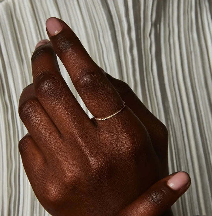 A close-up of a hand with dark skin, fingers gently touching one another, displaying a twisted stacking ring - 10k yellow gold by bluboho on the middle finger. The background features a white, pleated fabric. The nails are neatly manicured.