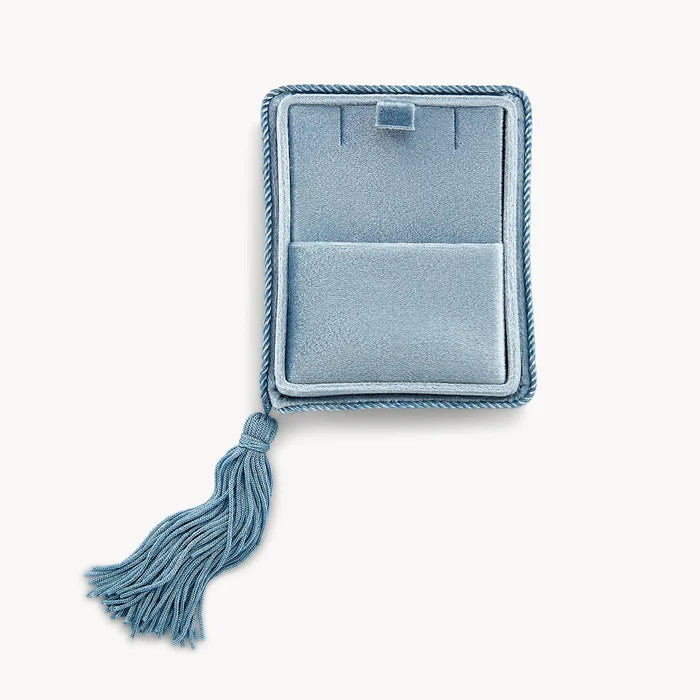 The velvet tassel box from bluboho lies open, its plush light blue velvet exterior adorned with a twisted rope border and a matching tassel. Inside, the box features a rectangular slot for securing a necklace and a bottom compartment for additional storage. The background is plain white.