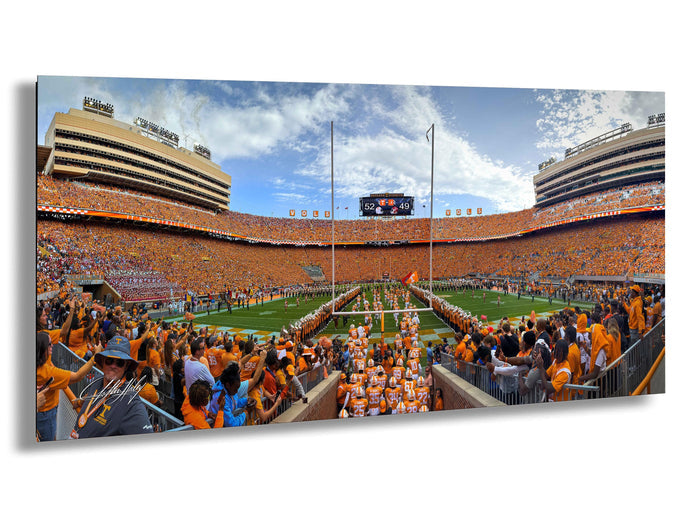 A large crowd of people wearing orange clothing gathered in a stadium, with a football field visible in the background.