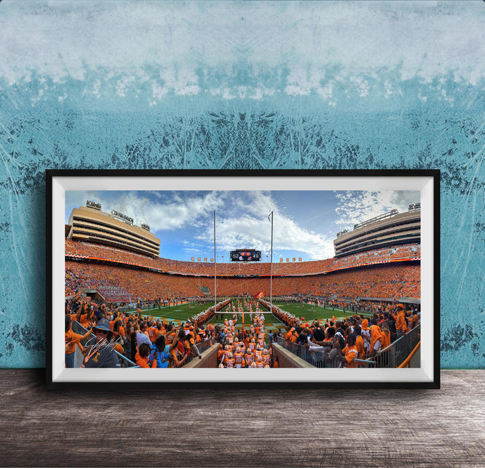 A framed photograph of a crowded football stadium with a large crowd of spectators, a large scoreboard, and a large field.