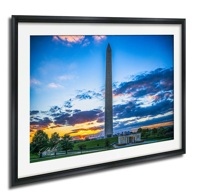 a tall obelisk against a colorful sunset sky, with a building and trees in the foreground.