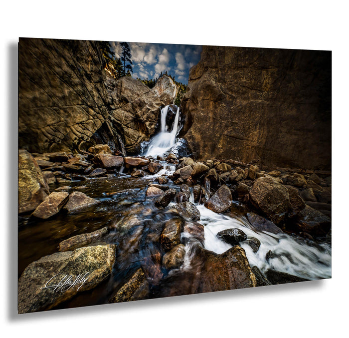 A serene waterfall cascades over rocks in a rocky canyon, surrounded by lush greenery and a cloudy sky.