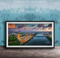 The image is a framed photograph of the Palace of Westminster, also known as the Houses of Parliament, in London, England. The photograph captures the iconic skyline of the city, with the River Thames flowing in the foreground. The sky is filled with vibrant colors, suggesting a beautiful sunset or sunrise. The photograph is framed and displayed on a wooden surface, creating a visually appealing and artistic composition.