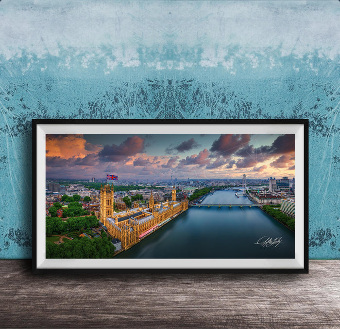 The image is a framed photograph of the Palace of Westminster, also known as the Houses of Parliament, in London, England. The photograph captures the iconic skyline of the city, with the River Thames flowing in the foreground. The sky is filled with vibrant colors, suggesting a beautiful sunset or sunrise. The photograph is framed and displayed on a wooden surface, creating a visually appealing and artistic composition.
