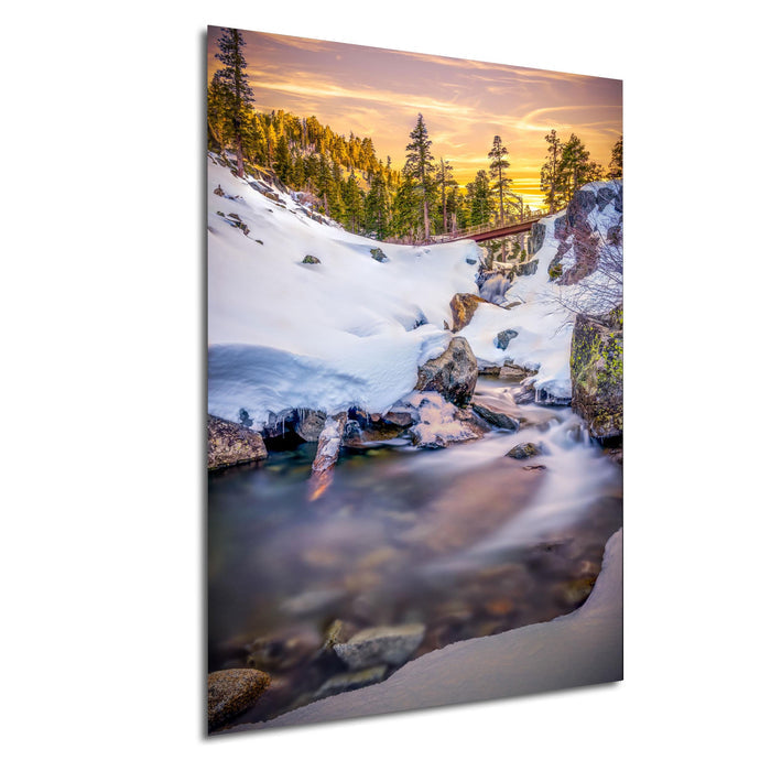 A serene winter landscape with a snow-covered mountain, a flowing stream, and a forest in the background, all bathed in the warm glow of a sunset.