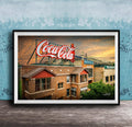 A framed photograph of a Coca-Cola sign on top of a brick building, with a sunset sky in the background.
