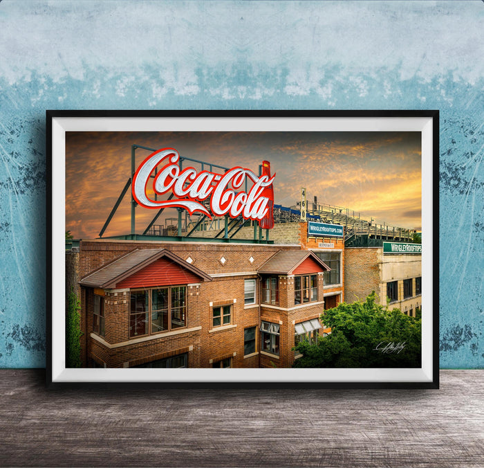 A framed photograph of a Coca-Cola sign on top of a brick building, with a sunset sky in the background.