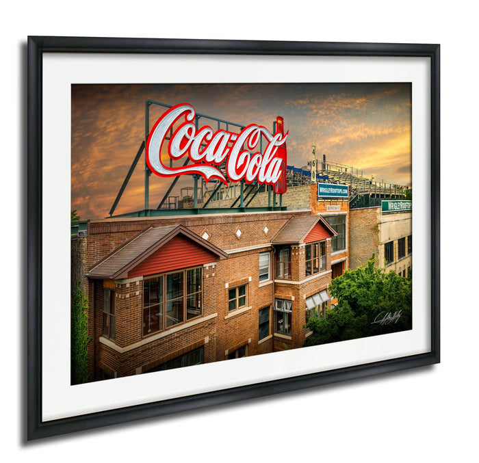 A framed image of a large Coca-Cola sign on top of a brick building, with a cloudy sky in the background.