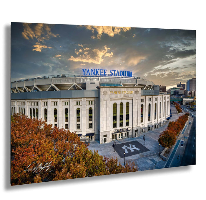 the Yankee Stadium, a large sports stadium located in New York City. The stadium is surrounded by trees with autumn foliage, and the sky is filled with clouds. The stadium is prominently displayed with the words "Yankees Stadium" written on it.