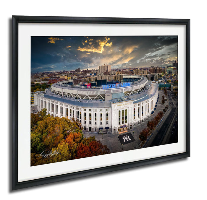 The image is a framed photograph of the Yankee Stadium, a large circular stadium located in New York City. The stadium is surrounded by a cityscape with buildings and trees, and the sky is filled with clouds. The photograph is framed in a black frame and is hung on a wall.