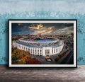 The image is a framed photograph of the New York Yankees stadium, which is a large, circular stadium located in the city of New York. The photograph captures the stadium's distinctive architecture and surrounding cityscape, with a dramatic sky in the background.