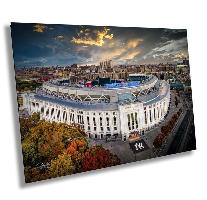 a large, circular stadium with a curved roof, surrounded by trees and buildings in the background. The sky above the stadium is cloudy, and the sun is setting, creating a dramatic and colorful atmosphere.