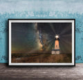 A framed photograph depicting a lighthouse standing in a field at night, with a starry sky and a galaxy in the background.