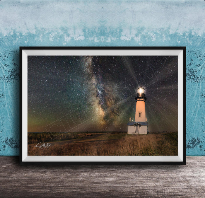 A framed photograph depicting a lighthouse standing in a field at night, with a starry sky and a galaxy in the background.