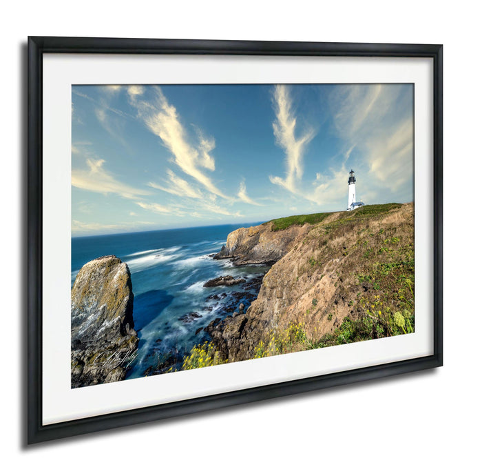 A framed photograph depicting a lighthouse on a cliff overlooking the ocean, with a cloudy blue sky in the background.