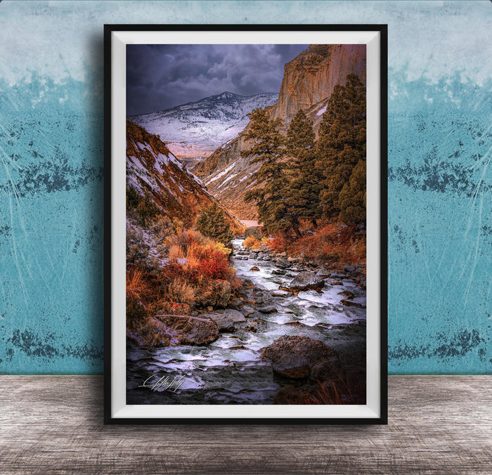 The image is a framed photograph of a scenic mountain landscape with a winding river flowing through it. The mountains are covered in snow, and the sky is filled with clouds, creating a dramatic and moody atmosphere. The photograph is framed and displayed on a wooden surface, with a blue wall in the background.